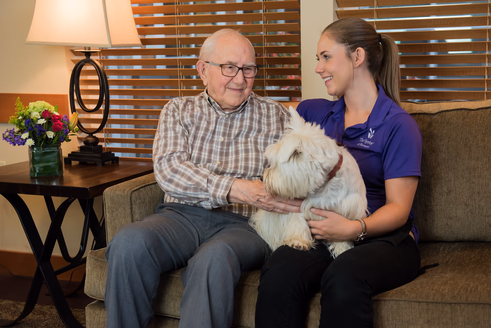 An elderly man and a young woman sitting on a couch indoors. The elderly man is wearing glasses and a plaid shirt, smiling while petting a small white dog sitting on the woman's lap. The woman, wearing a purple polo shirt with The Springs at Sherwood logo, is smiling at the man. A side table with a lamp and a vase of flowers is visible next to the couch.