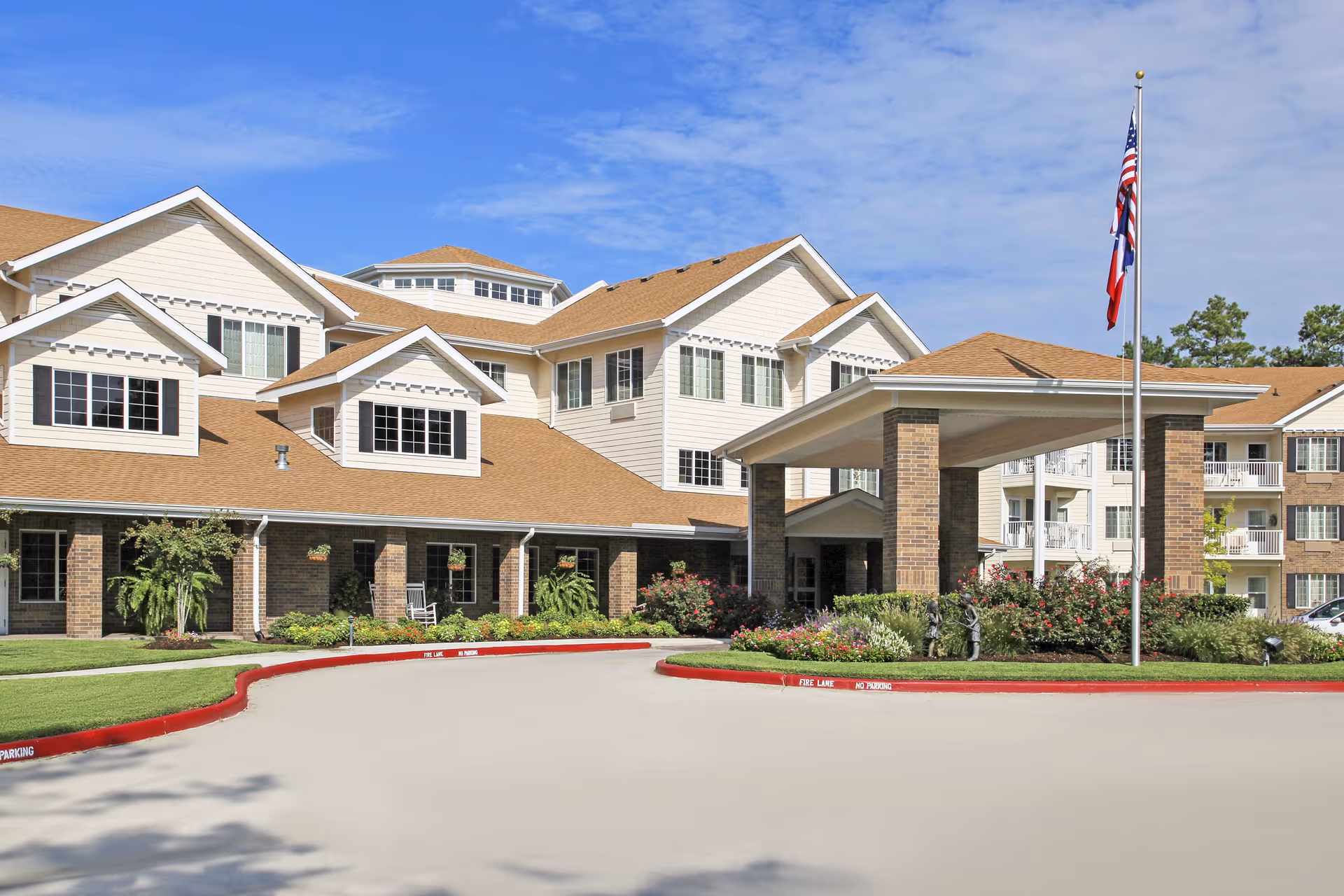 Exterior view of a multi-story senior living facility with beige siding and brown roofs. The building features multiple windows, a covered entrance supported by brick columns, landscaped greenery, and an American flag on a flagpole in front.