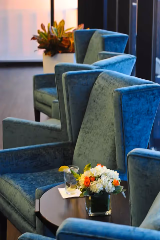 A cozy seating area with blue upholstered armchairs arranged around a dark wooden table. On the table, there is a glass of water with a lemon slice on the rim and a small floral arrangement with white and orange flowers. In the background, there is a large window and a potted plant.