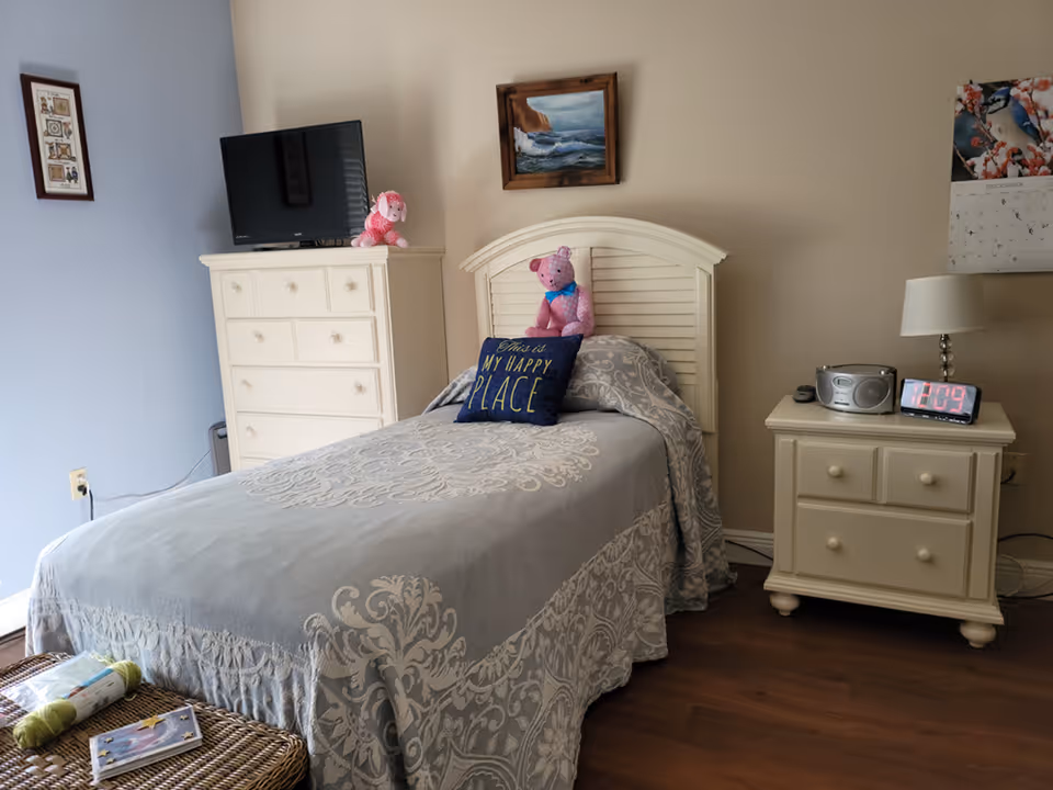 A tidy single bedroom with a twin bed, white headboard and furniture, a TV on a dresser, stuffed animals and a nightstand with a lamp and digital clock.
