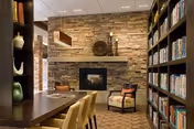 A cozy interior room featuring a stone fireplace with decorative items on the mantel, two upholstered chairs nearby, a long wooden table with chairs, and bookshelves filled with books on the right side.