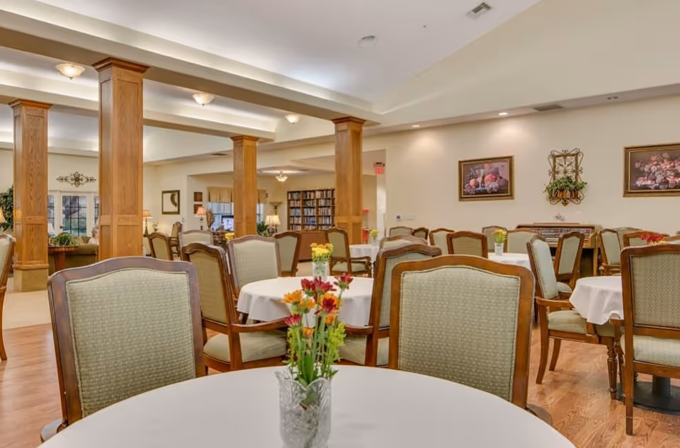 Communal dining room with round tables covered in white tablecloths, wooden chairs, floral centerpieces, and decorative wooden columns.