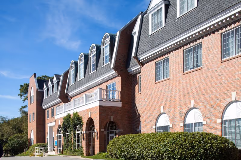 Front exterior of a red brick building with arched windows, dormer windows, a small balcony, and landscaped shrubs under a blue sky.