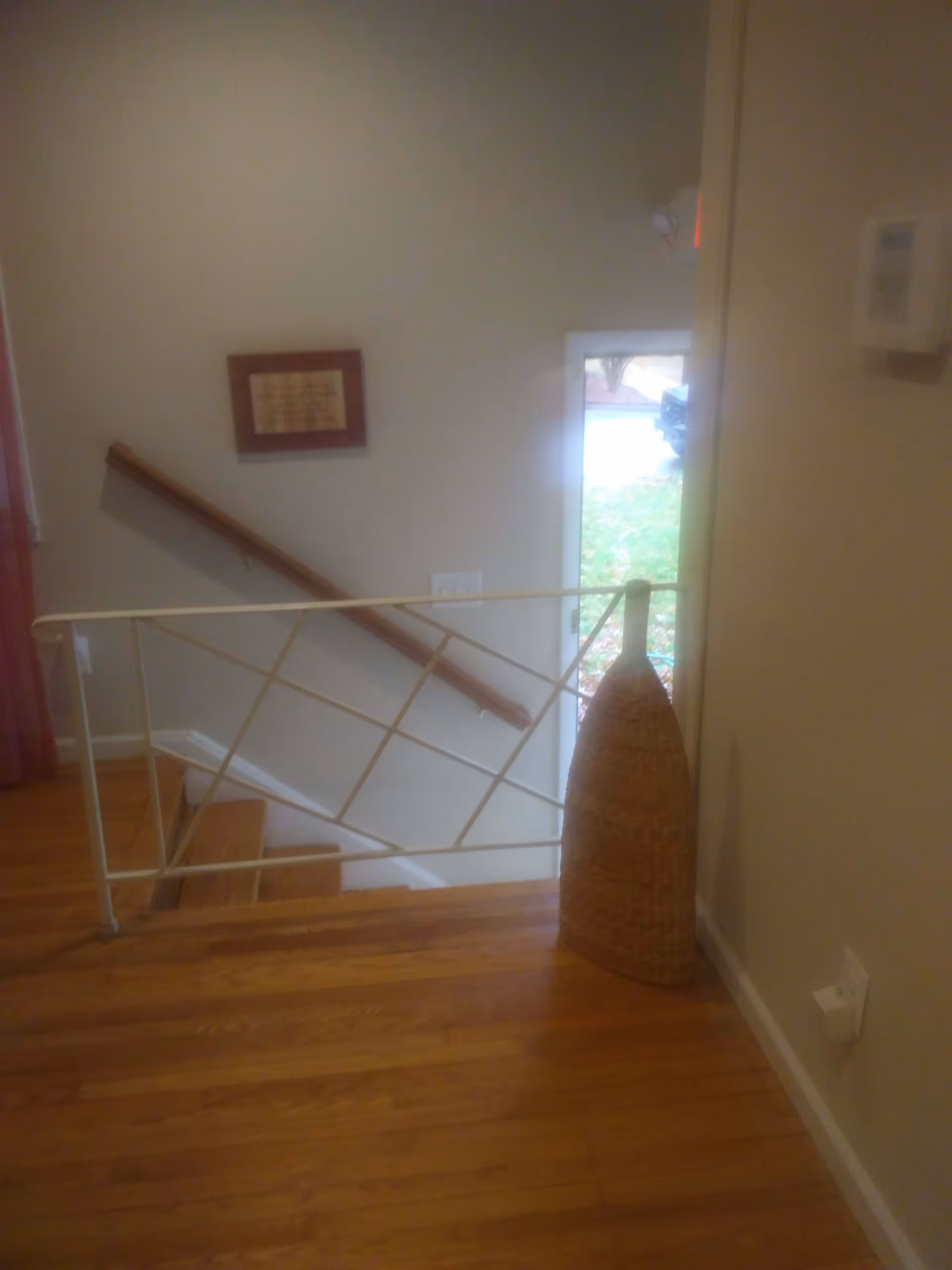 Interior view of a stairway area with wooden steps and a metal railing. A large woven basket is placed on the wooden floor near the wall. A framed picture hangs on the wall above the handrail, and a door with a window shows a glimpse of the outside greenery.