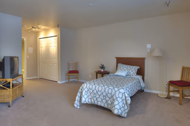 A simple bedroom in an assisted living facility featuring a single bed with patterned bedding, two wooden chairs with red cushions, a small round table with a flower arrangement, a floor lamp, and a TV on a wicker stand. The room has beige walls and carpeted floor, with a closet and hallway visible in the background.