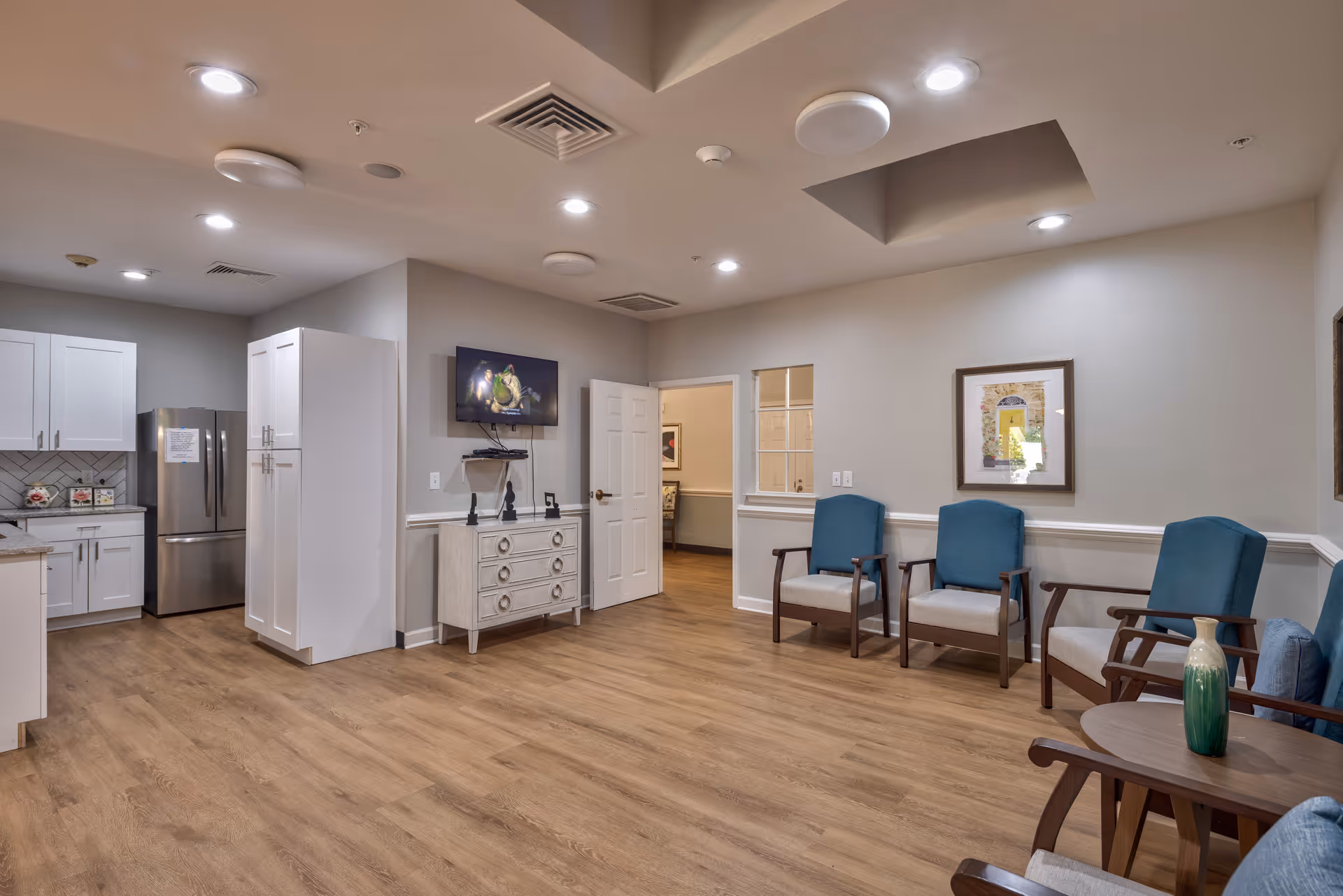 A spacious room with wood flooring featuring a small kitchen area with white cabinets and a stainless steel refrigerator on the left. On the right side, there are four blue cushioned chairs arranged along the wall with a small round table holding a green vase. A flat-screen TV is mounted on the wall above a white dresser. The walls are painted light gray with white trim, and there is a framed picture hanging above the chairs. The ceiling has recessed lighting and ventilation fixtures.