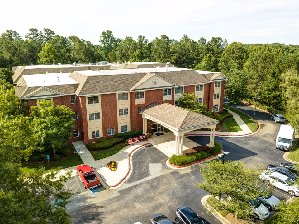 Aerial view of Magnolia Place of Roswell, a three-story brick building with a covered entrance and a driveway. Several cars are parked in the parking lot surrounding the building, and the area is surrounded by green trees and landscaping.