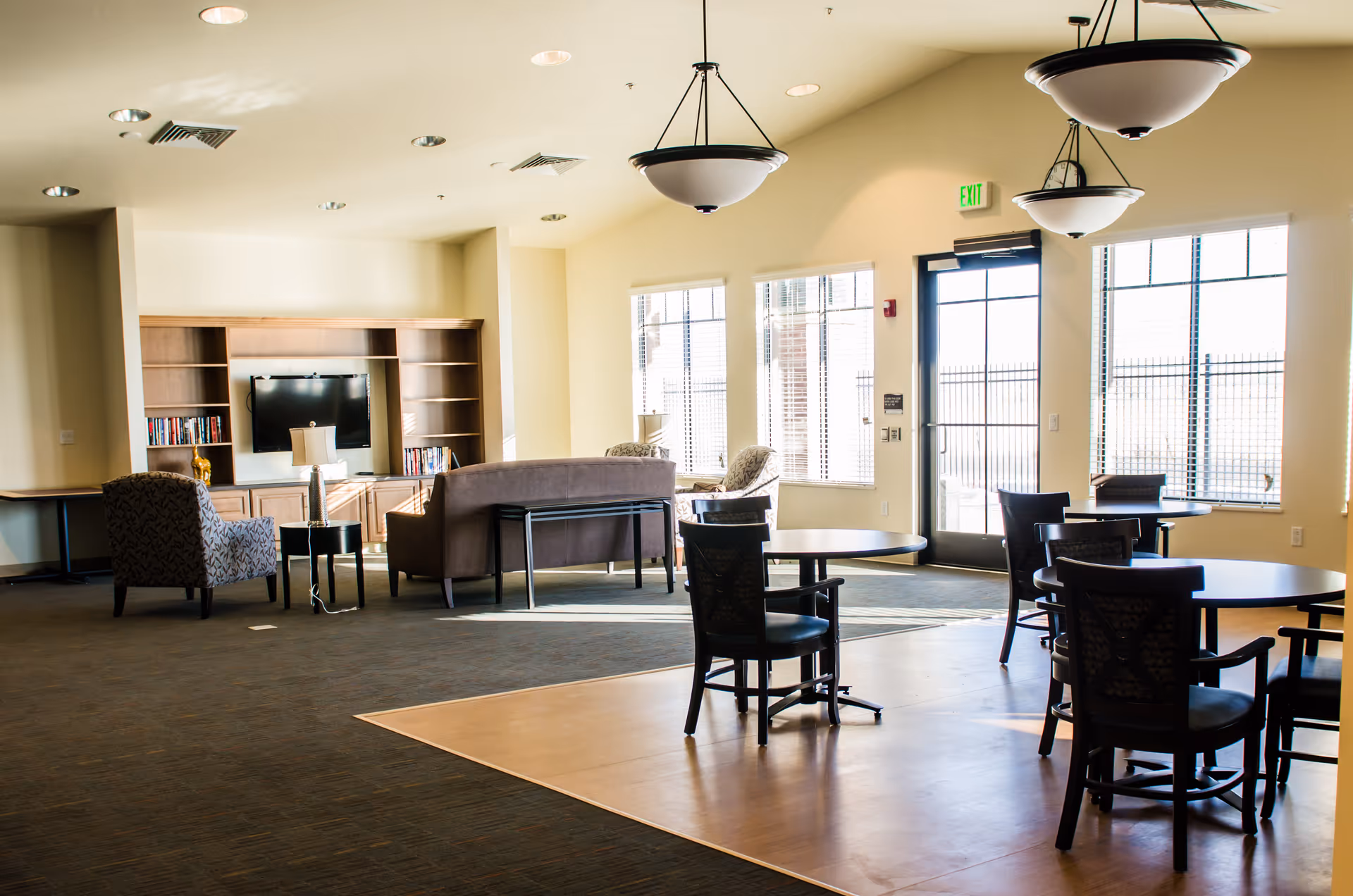 A bright and spacious common area in a senior living facility with several round tables and chairs on a wooden floor section. In the background, there is a seating area with a sofa, armchairs, a TV mounted on a wooden entertainment center, and large windows letting in natural light. Ceiling lights hang from the high ceiling.