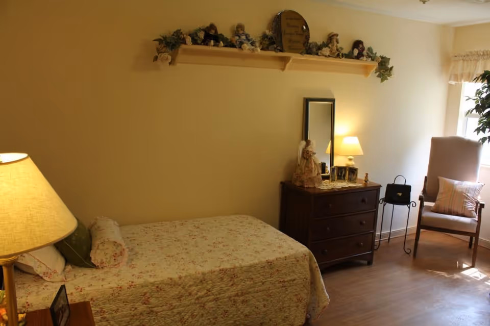 A cozy bedroom with a single bed covered in a floral quilt, a wooden nightstand with a lamp, a wooden dresser with a small lamp, a mirror, and decorative dolls on top. A cushioned chair with a pillow is placed near a window with light curtains, and a plant is visible in the corner.
