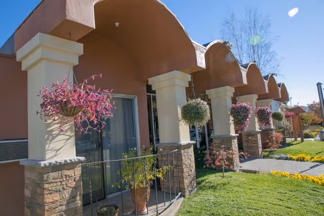 Exterior view of a building with arched roof sections and stone pillars. Hanging flower baskets and potted plants decorate the front porch area. The lawn is green and well-maintained with yellow flowers along the walkway under a clear blue sky.