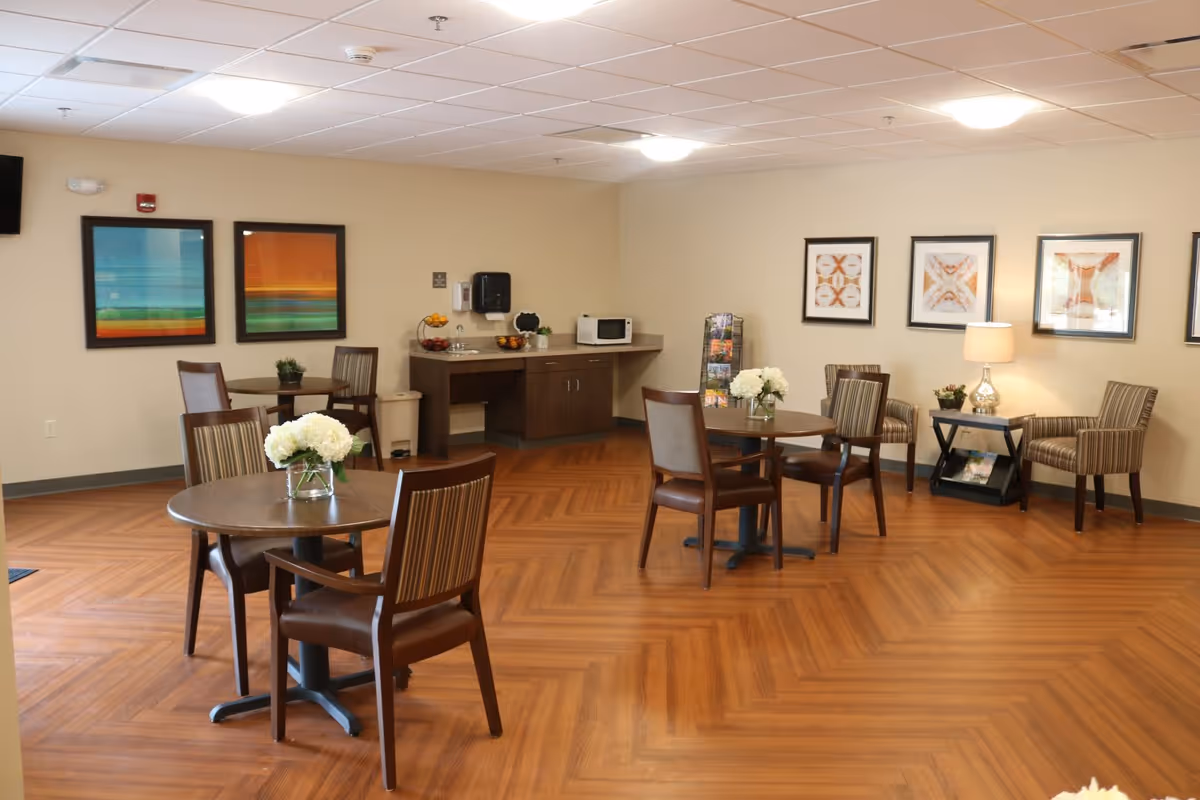 A senior living facility common area with wooden floors and beige walls. The room contains round tables with chairs, each table decorated with a vase of white flowers. There is a small kitchenette area with a microwave, sink, and fruit bowls. The walls are adorned with framed abstract artwork, and there is a side table with a lamp and additional chairs arranged nearby.