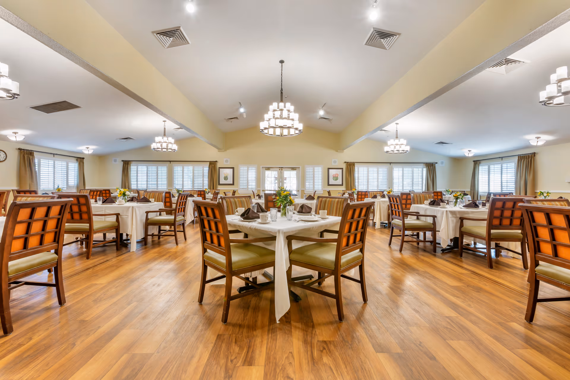A spacious dining room with wooden floors and multiple tables covered with white tablecloths. Each table is set with cups, glasses, plates, and brown folded napkins. The room is well-lit with modern chandeliers hanging from the ceiling and has large windows with white shutters and beige curtains. There are framed pictures on the walls and floral centerpieces on each table.