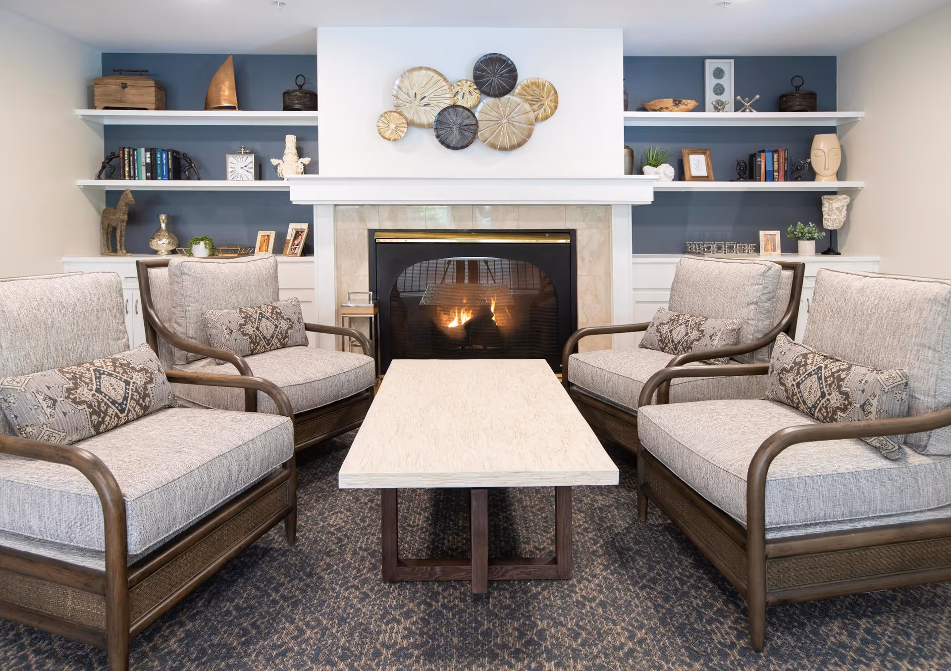 A cozy living room area with four cushioned armchairs arranged around a rectangular coffee table. Behind the seating is a fireplace with a fire burning, flanked by built-in shelves decorated with books, plants, and various decorative items. The walls are painted in neutral tones with blue accent panels behind the shelves.