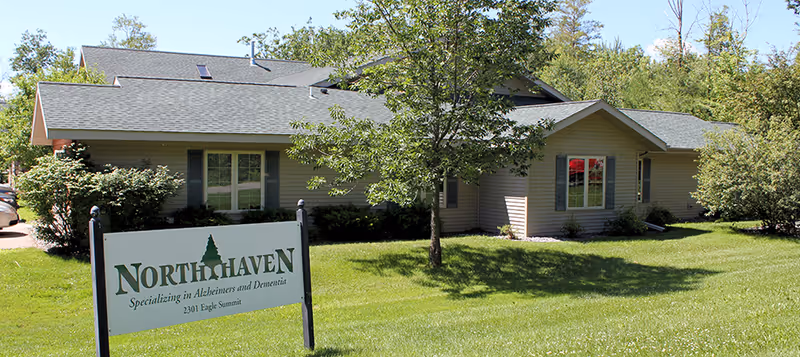 Exterior view of a single-story building with beige siding and a gray roof, surrounded by green grass, trees, and bushes. A sign in front reads 'North Haven Specializing in Alzheimers and Dementia 2301 Eagle Summit.'