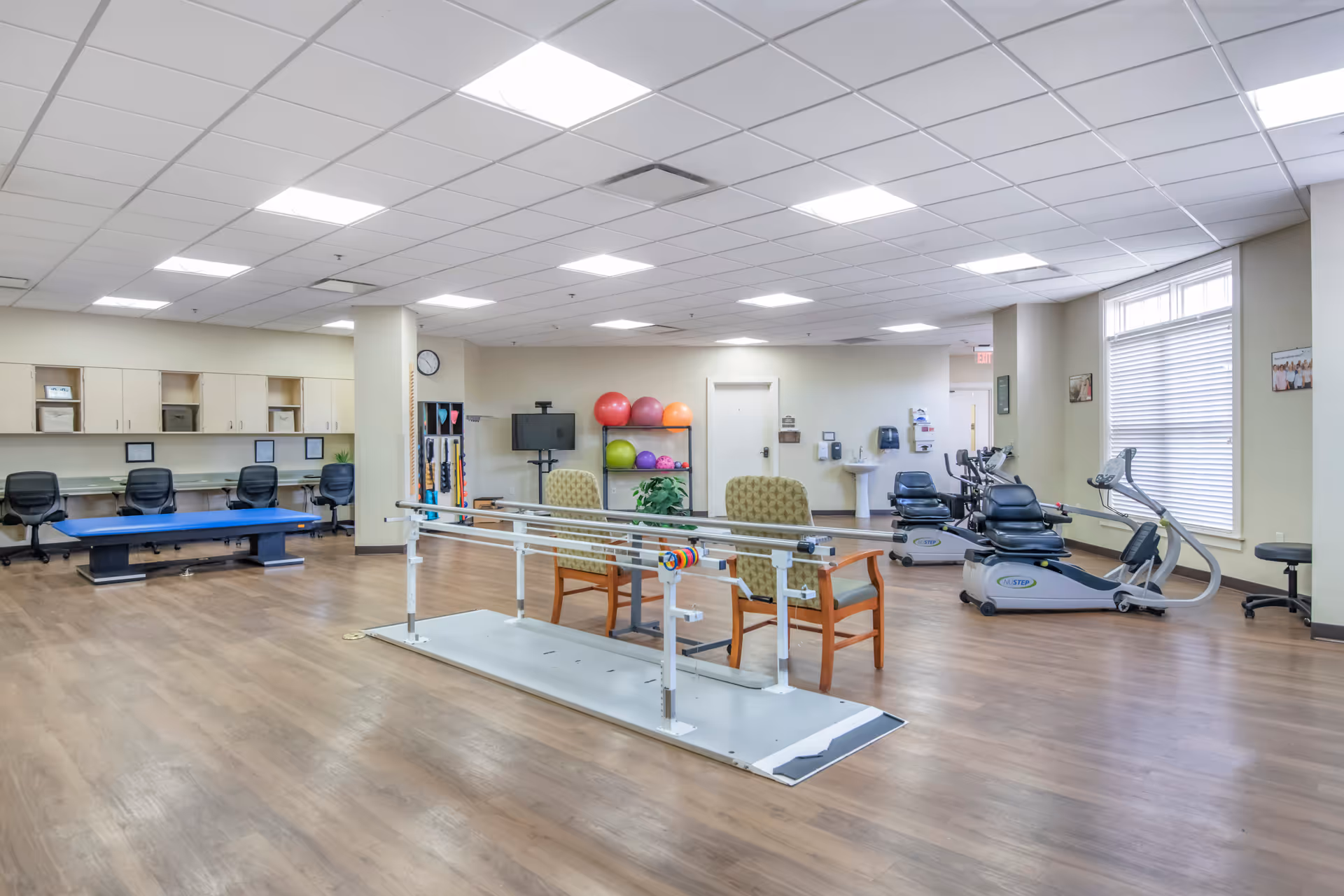 A spacious physical therapy or rehabilitation room with wooden flooring, exercise equipment including stationary bikes, parallel bars for walking practice, therapy balls on a rack, chairs, and a blue therapy table. The room has a drop ceiling with fluorescent lights and a large window with blinds letting in natural light.