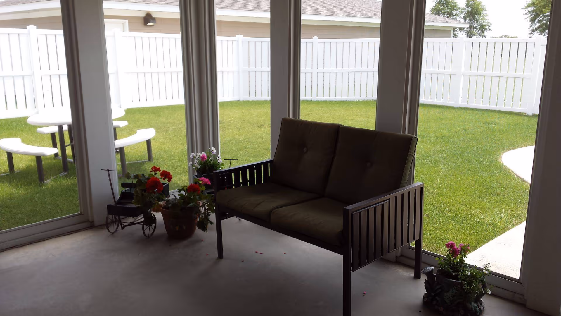 Indoor seating area with a cushioned bench and several potted plants near large glass windows overlooking a green lawn enclosed by a white fence. Outside, there is a white picnic table with benches on the grass.