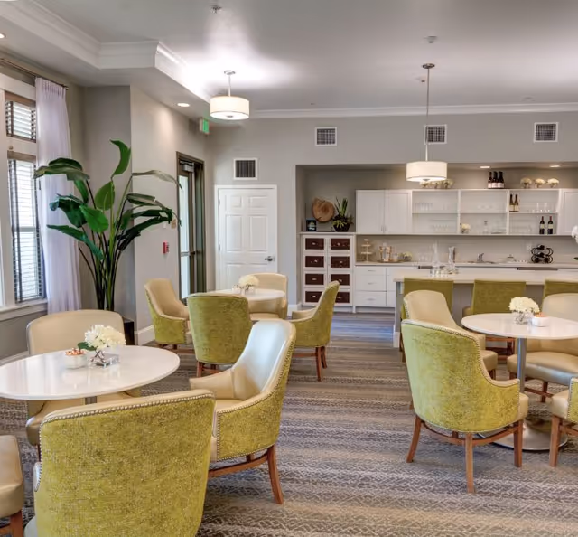 Bright communal dining/lounge area with round tables, green upholstered chairs, and a small kitchen/bar in the back.