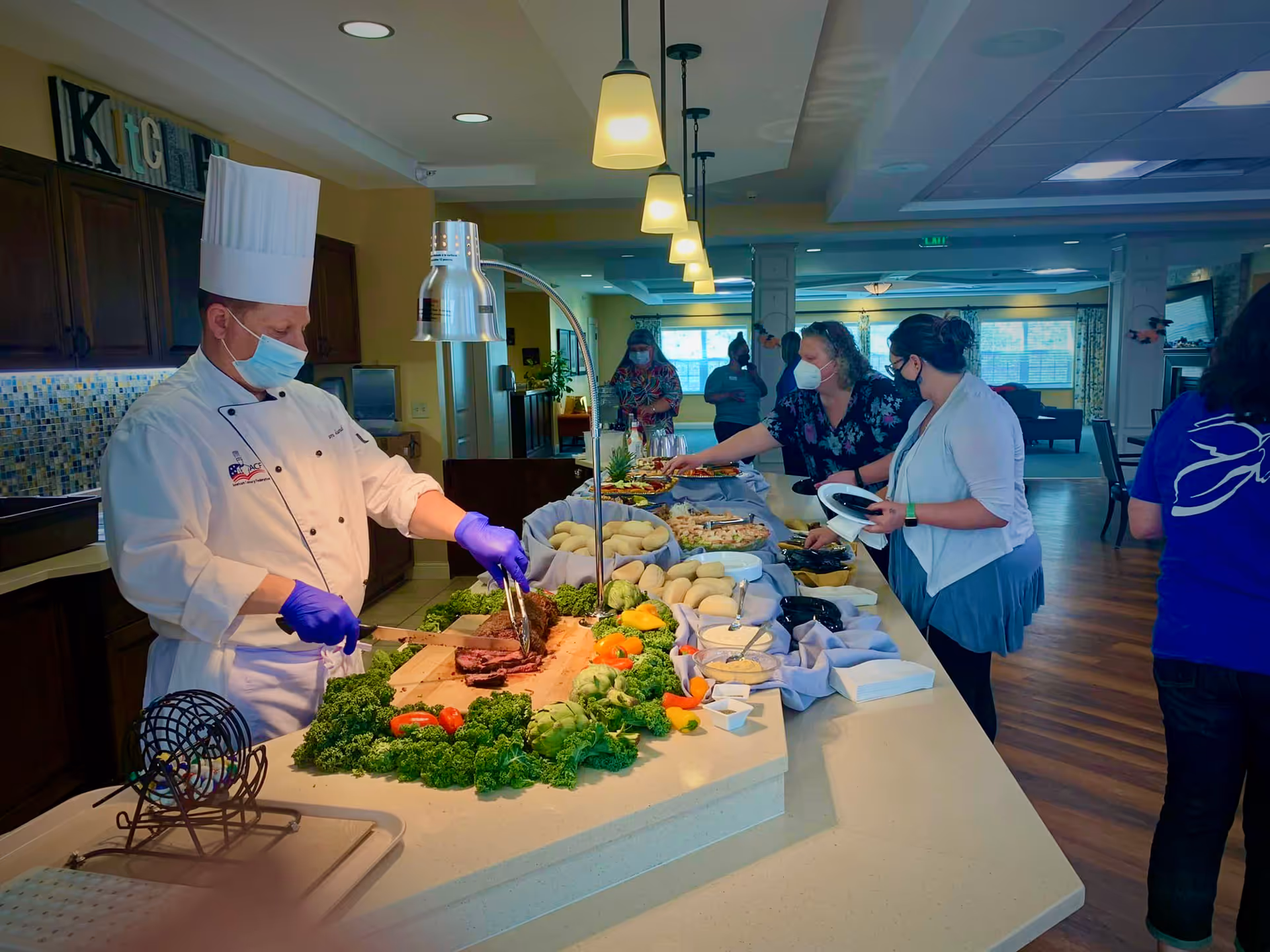 A chef wearing a white uniform, chef hat, face mask, and purple gloves is slicing meat on a counter decorated with leafy greens and vegetables. Several people wearing face masks are serving themselves food from a buffet-style setup in a well-lit kitchen or dining area with wooden floors and overhead pendant lights.