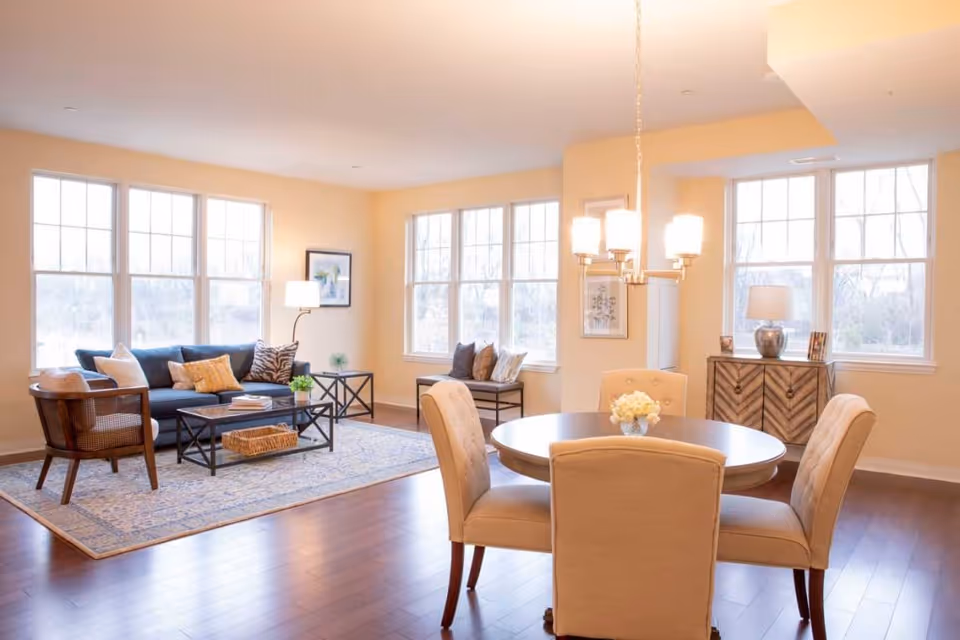 Bright and spacious living and dining area with large windows letting in natural light. The living room features a blue sofa with assorted pillows, a wooden chair, a coffee table with a basket underneath, and a side table with a lamp and plant. The dining area has a round table with four beige upholstered chairs and a chandelier overhead. A wooden sideboard with a lamp and framed photos is against the wall.