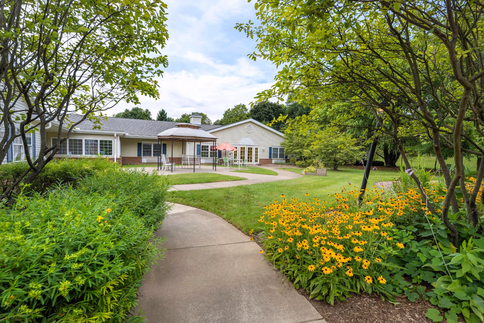 A paved walkway curves through a garden with green shrubs and yellow flowers leading to a single-story building with beige siding and multiple windows. There are outdoor tables with umbrellas on a patio area in front of the building, surrounded by trees and greenery under a partly cloudy sky.