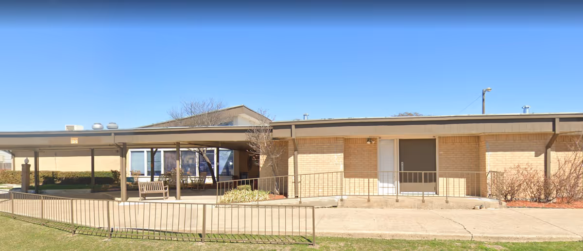 Single-story building with beige brick exterior and a covered patio area featuring benches and a table. There is a metal railing along the sidewalk and some sparse landscaping with bushes and a small tree. The sky is clear and blue.