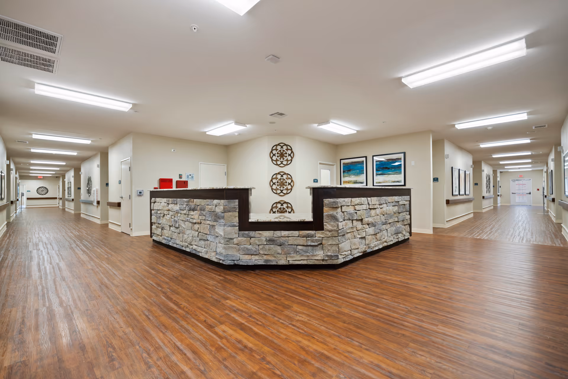 Interior view of a senior living facility hallway with a central stone reception desk. The hallway has wood flooring, beige walls, ceiling lights, and decorative wall art. Doors line both sides of the hallway.