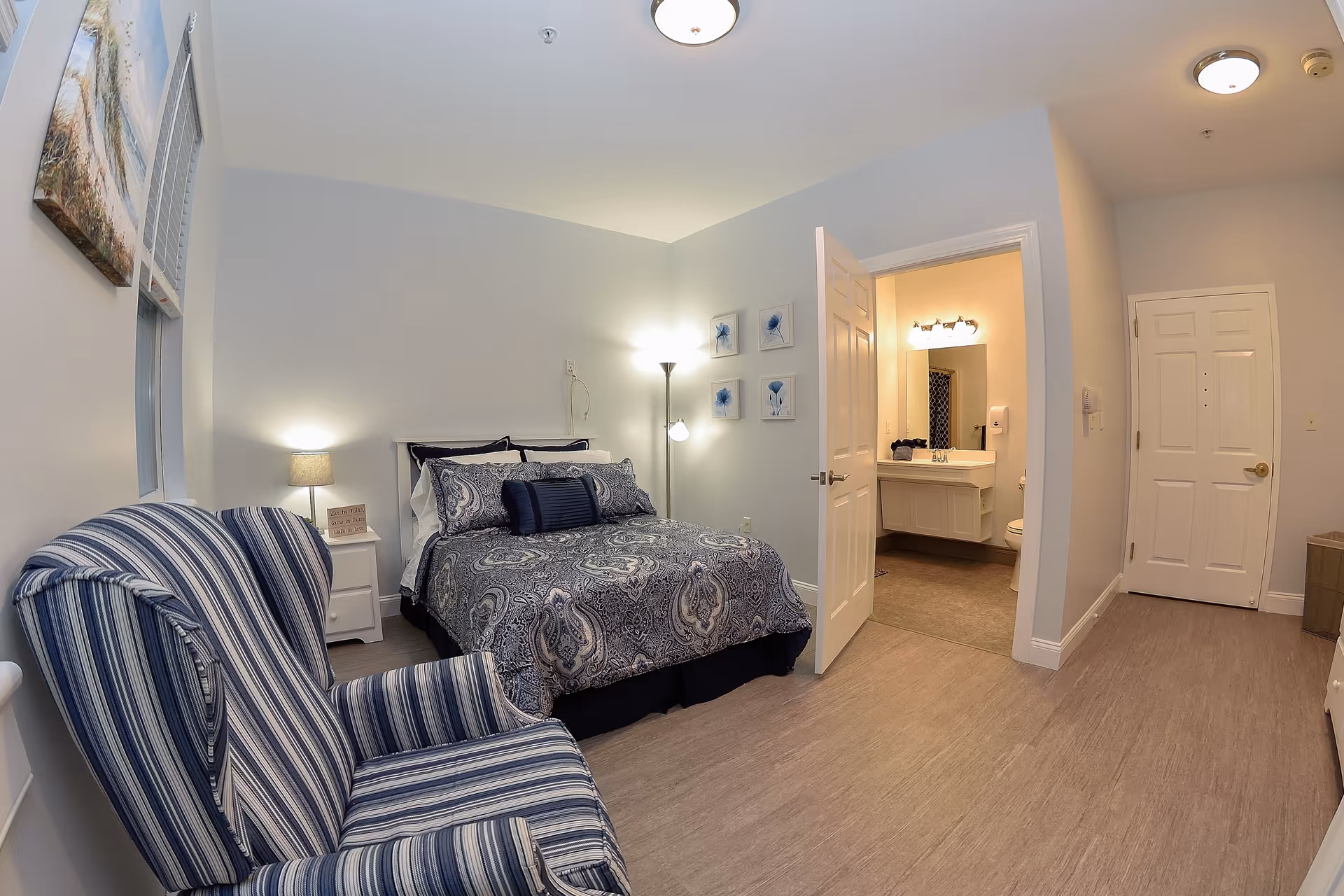 A cozy bedroom with a blue and white patterned bedspread on a double bed, a striped blue armchair, a small white nightstand with a lamp, and a door leading to a bathroom with a sink and mirror. The walls are light-colored and decorated with blue floral artwork.