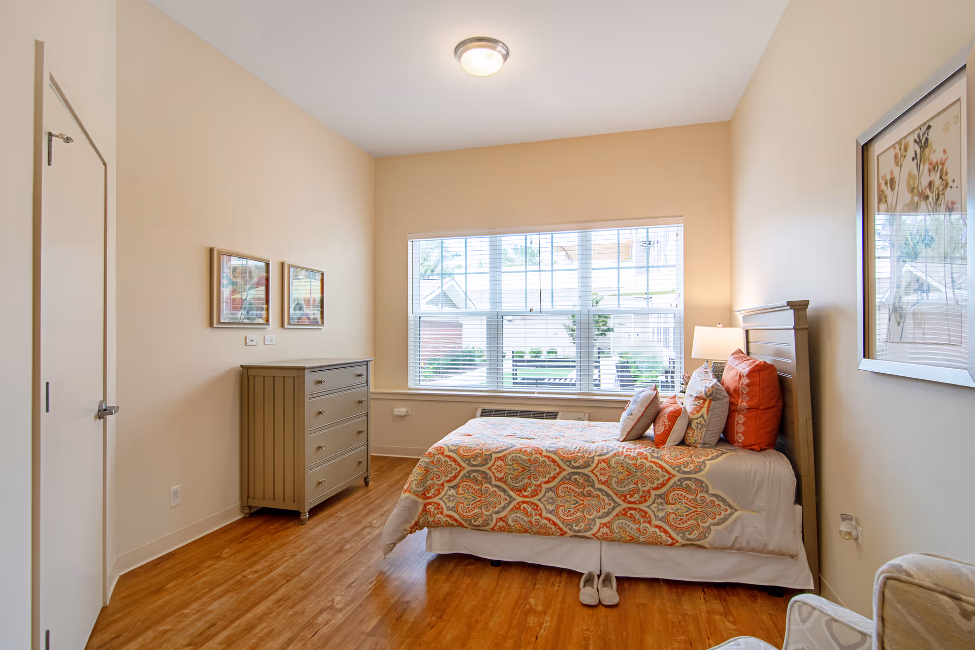 Sunny furnished bedroom with a single bed with patterned bedding, a dresser, window with blinds, and wood flooring.