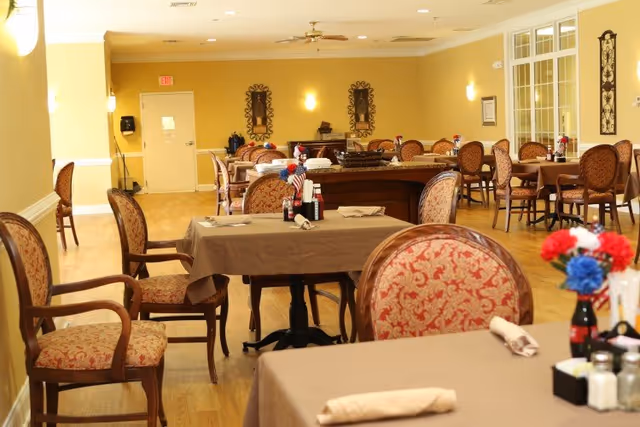 A dining room with multiple tables covered in beige tablecloths, each set with napkins and condiments. The chairs have wooden frames with patterned upholstery. The room has yellow walls, wooden flooring, wall sconces, and decorative wall hangings. There is a door at the far end and windows on the right side.