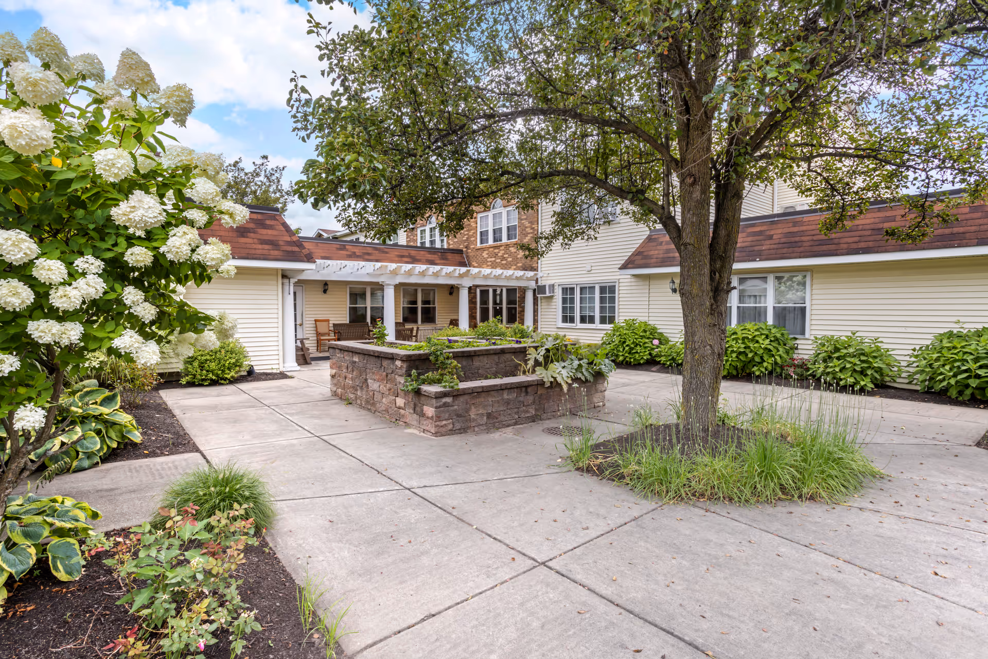 Outdoor courtyard area at Brookdale Castle Gardens featuring a large tree surrounded by grass, a raised stone planter with greenery, white flowering bushes, and a building with beige siding and brown roof shingles in the background.
