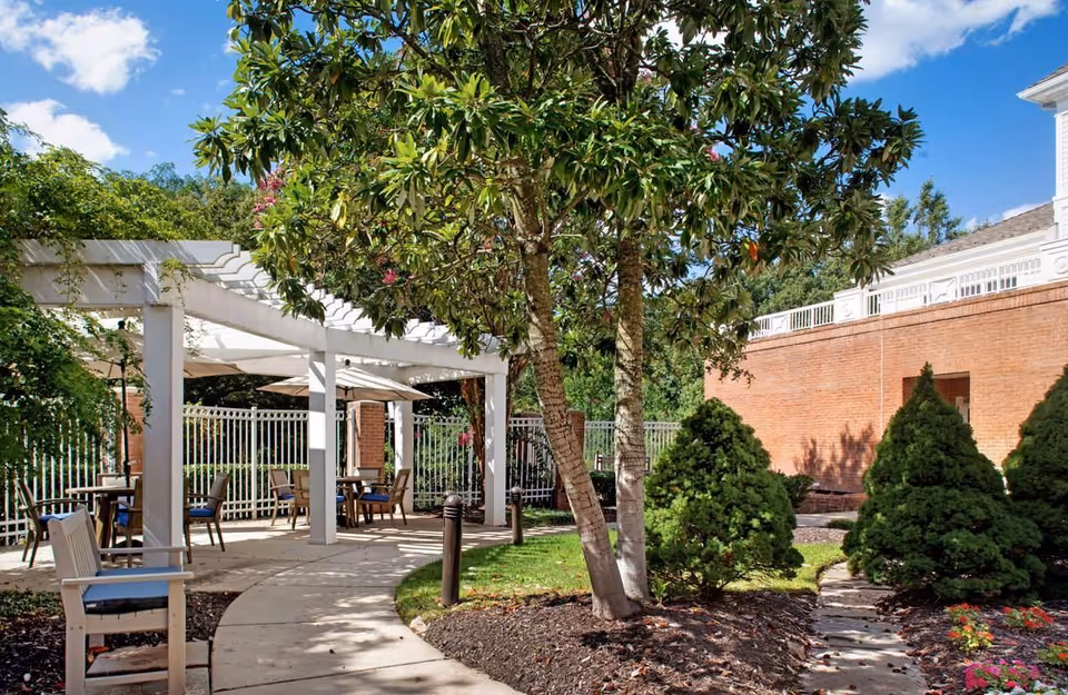 Outdoor garden area with a curved concrete pathway, green grass, trees, and shrubs. There is a white pergola with tables and chairs underneath, and a brick building wall in the background under a blue sky with some clouds.