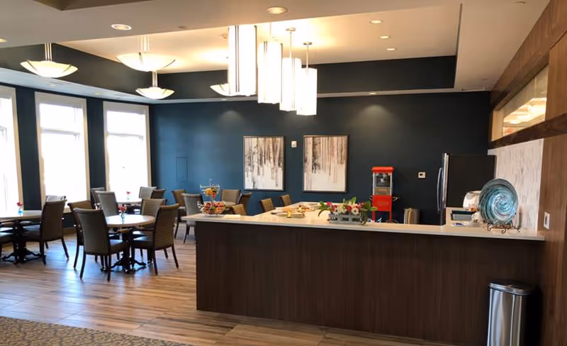 Bright communal dining area with tables and chairs, a long service counter topped with pendant lights and decorative accents, and artwork on a dark accent wall.