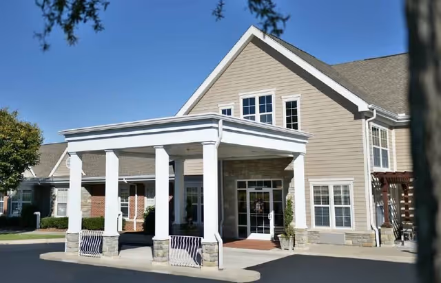 Front entrance of a two-story beige senior living building with a covered porte-cochère and glass double doors.