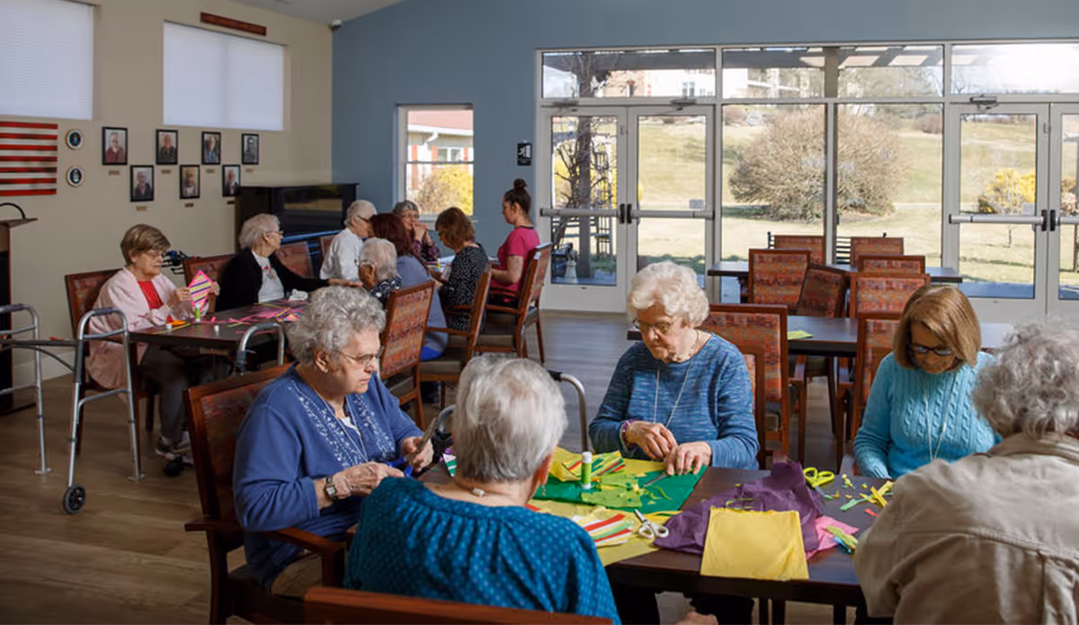 A group of elderly women sitting around tables in a bright room with large windows, engaging in arts and crafts activities with colorful paper and scissors. The room has wooden floors, several chairs, and tables, with a view of a grassy outdoor area through the windows.