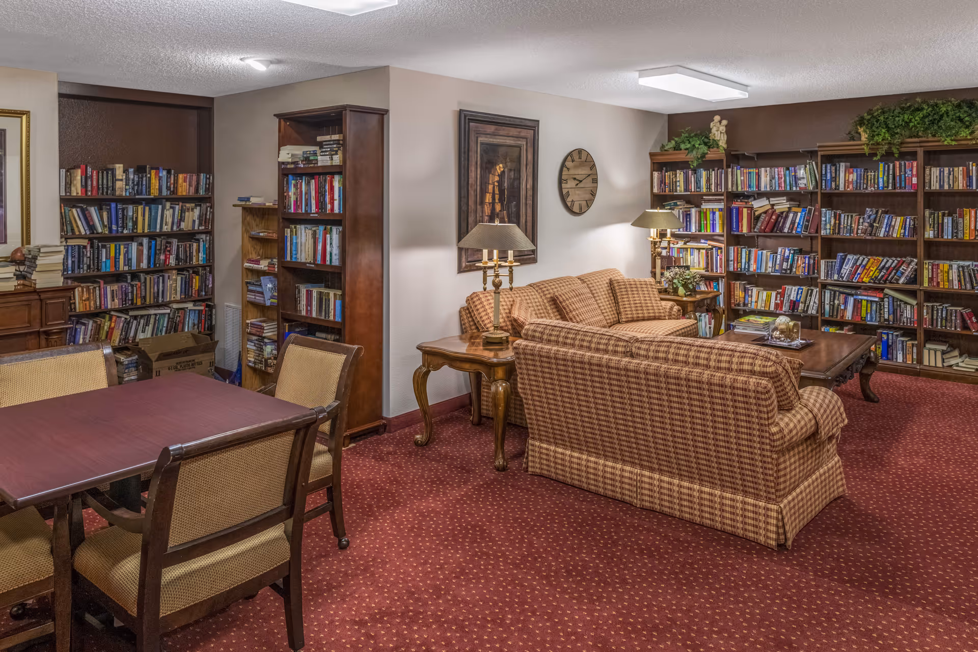 Cozy communal library and lounge with bookshelves lining the walls, upholstered sofas, lamps, and a table with chairs.