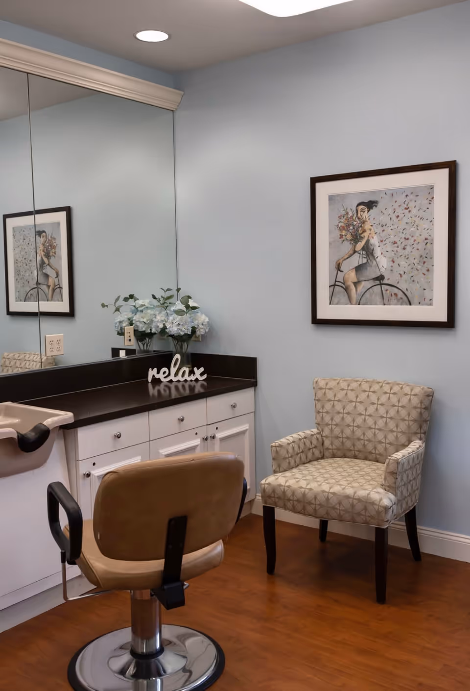 A small salon or grooming area with a beige salon chair in front of a counter with white cabinets and a large mirror. On the counter, there is a vase with white flowers and a decorative sign that says 'relax'. To the right, there is a patterned armchair and a framed artwork of a woman holding flowers while sitting on a bicycle, hanging on a light blue wall. The floor is wooden.