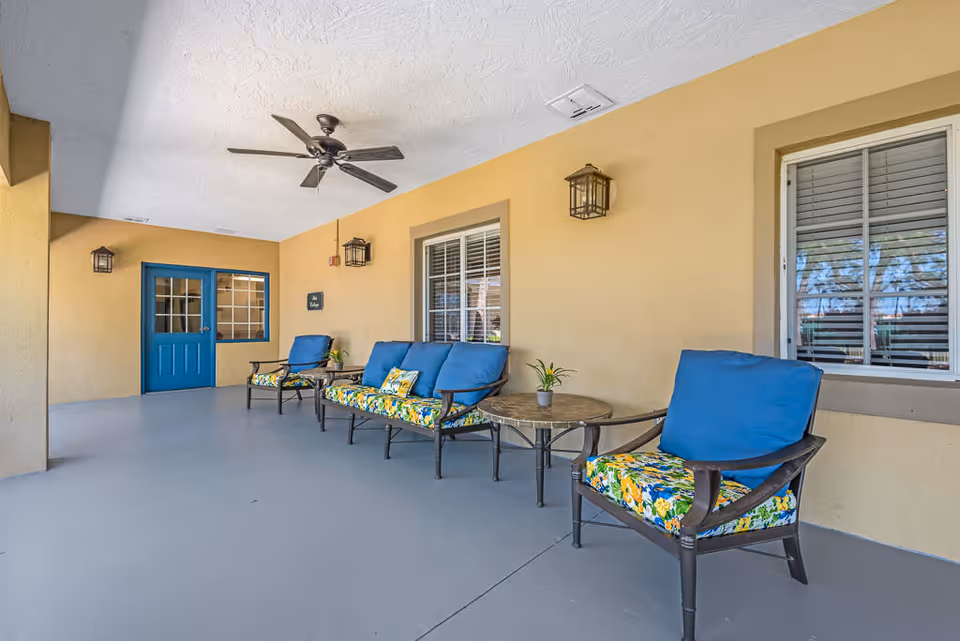 Covered outdoor patio area with beige walls and ceiling, featuring a ceiling fan, three cushioned chairs with blue backs and floral seat cushions, two small round tables with potted plants, and a blue door with windows in the background.