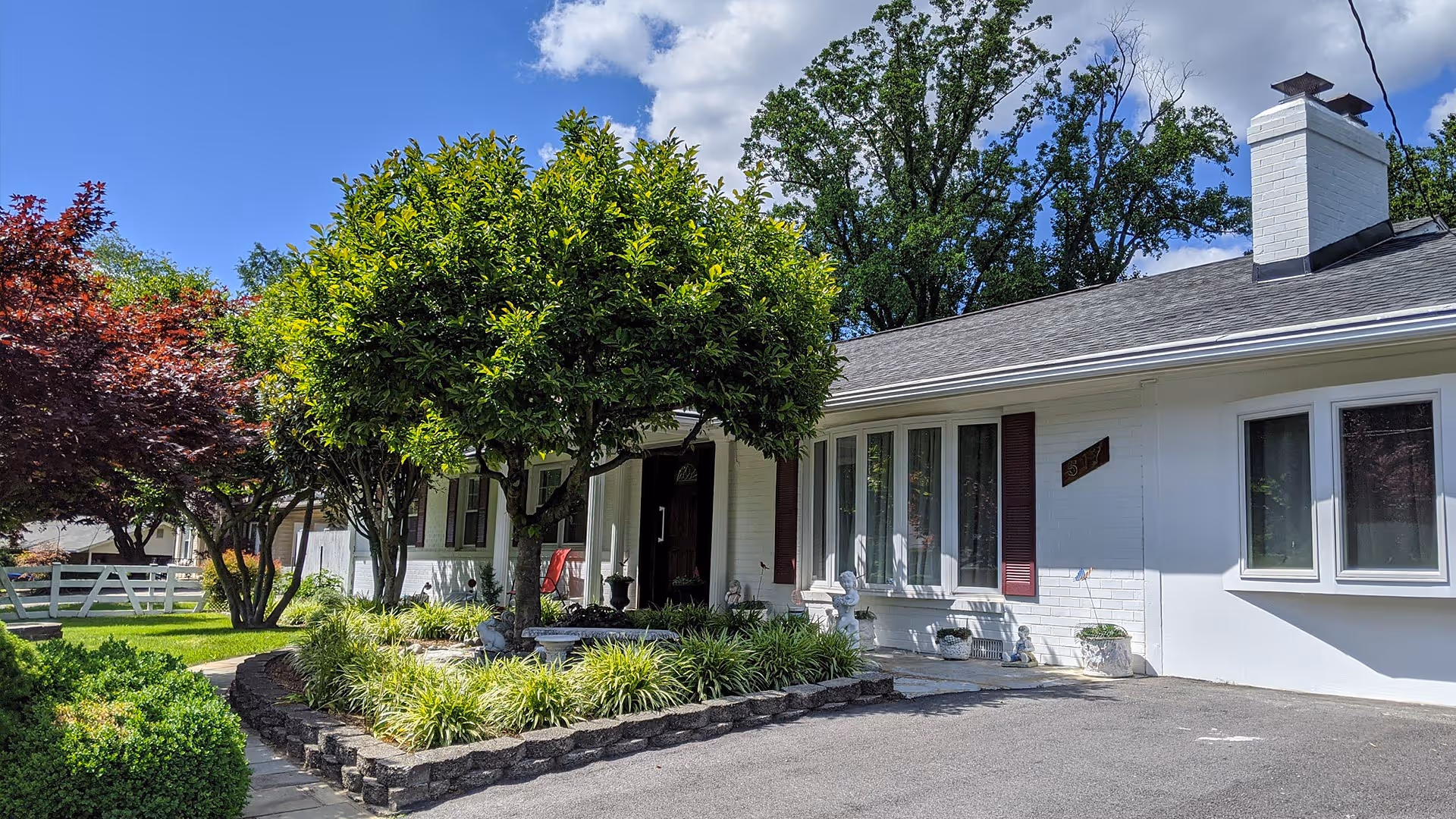 Exterior view of a single-story white brick building with a dark shingled roof, surrounded by green trees and shrubs under a blue sky with some clouds. There is a paved driveway in front and a small garden area with a tree near the entrance.