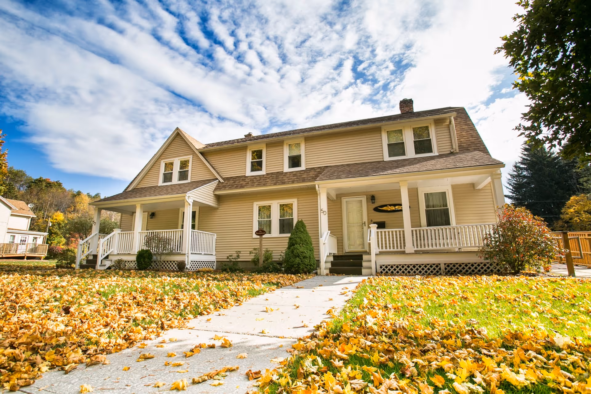 Two-story beige house with a front porch and a sidewalk through a yard covered in autumn leaves under a partly cloudy sky.