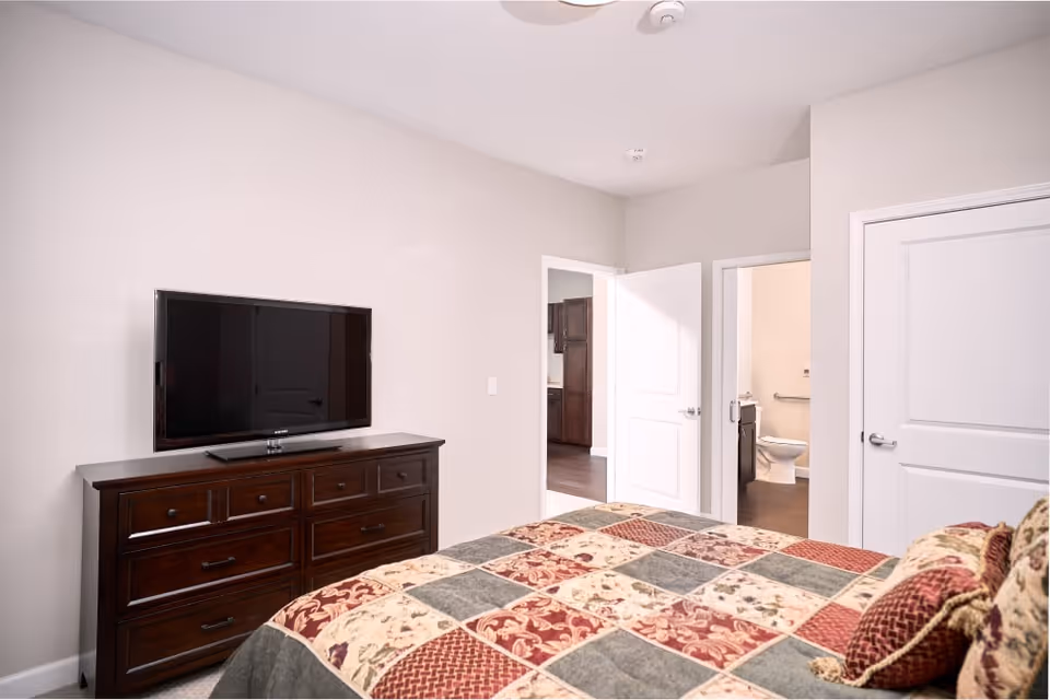 Bedroom with a patterned quilted bed, a dark wood dresser topped by a flat-screen TV, and open doors leading to a bathroom and hallway.