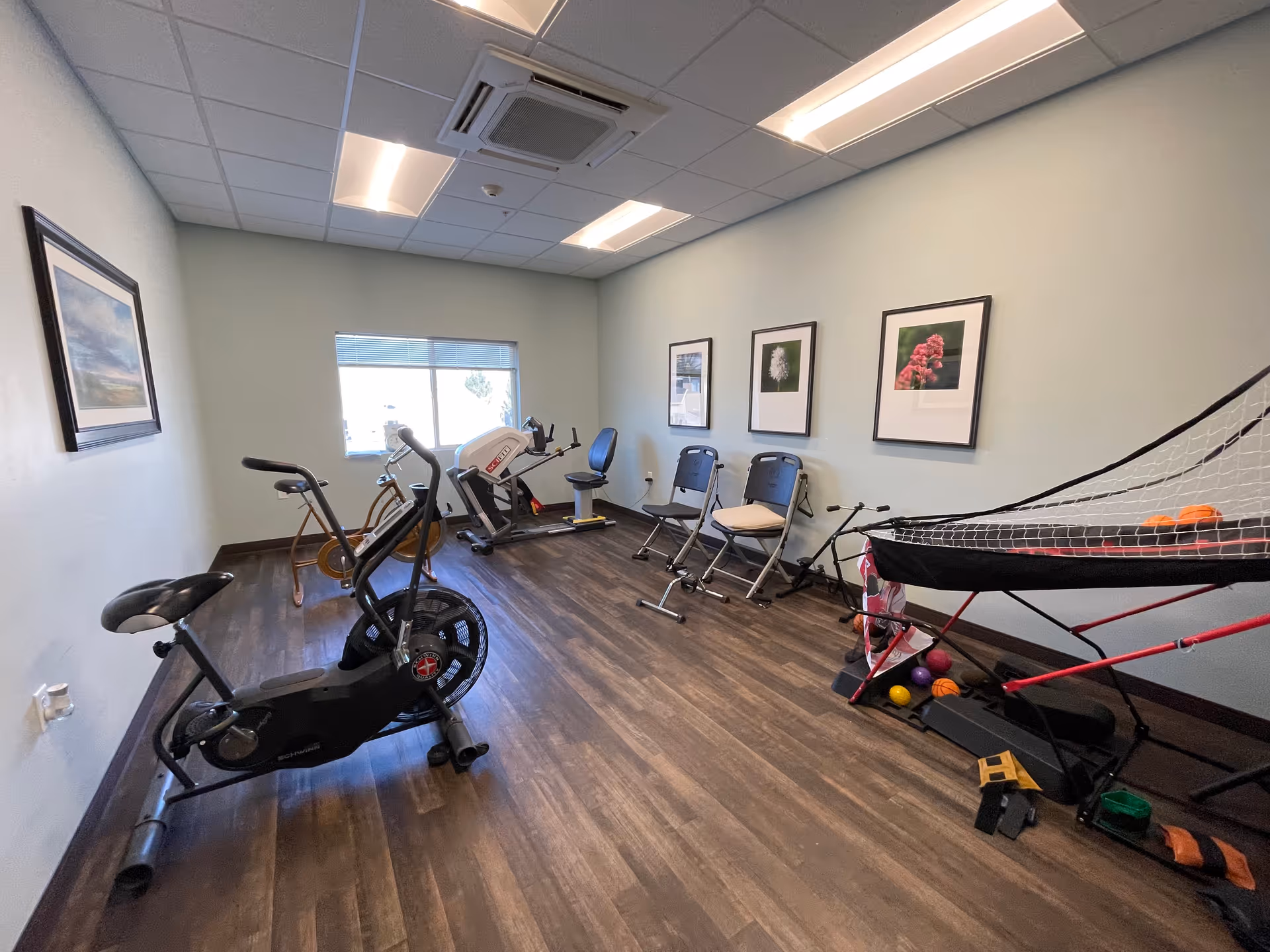 A small exercise room with wooden flooring and light-colored walls. The room contains various exercise equipment including a stationary bike, a recumbent bike, and a seated elliptical machine. There are three folding chairs lined up against the wall beneath three framed pictures of flowers. On the right side, there is a basketball arcade game with several small balls on the floor nearby. A window lets in natural light, and the ceiling has recessed lighting and an air conditioning unit.