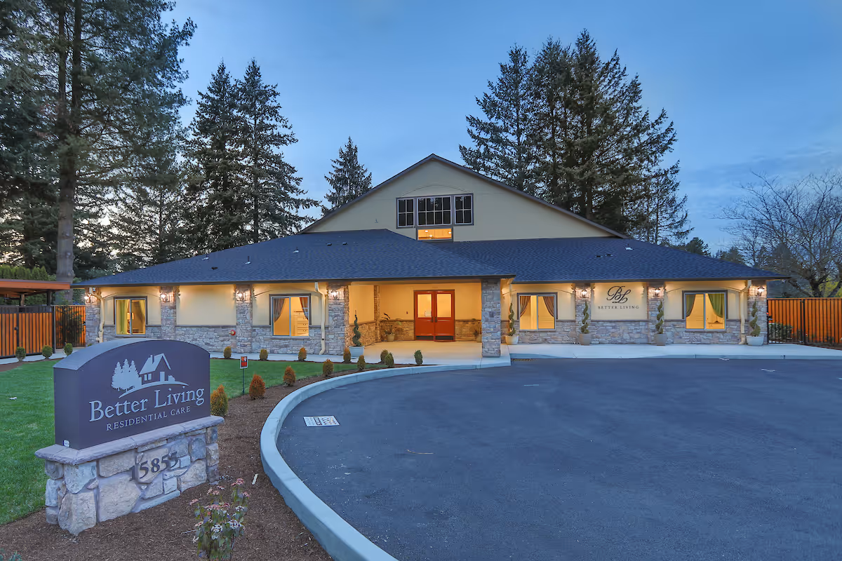 Exterior view of Better Living Residential Care facility at dusk, showing a single-story building with stone and beige siding, lit windows, and a curved driveway in front. There is a sign in the foreground with the facility name and address number 585.