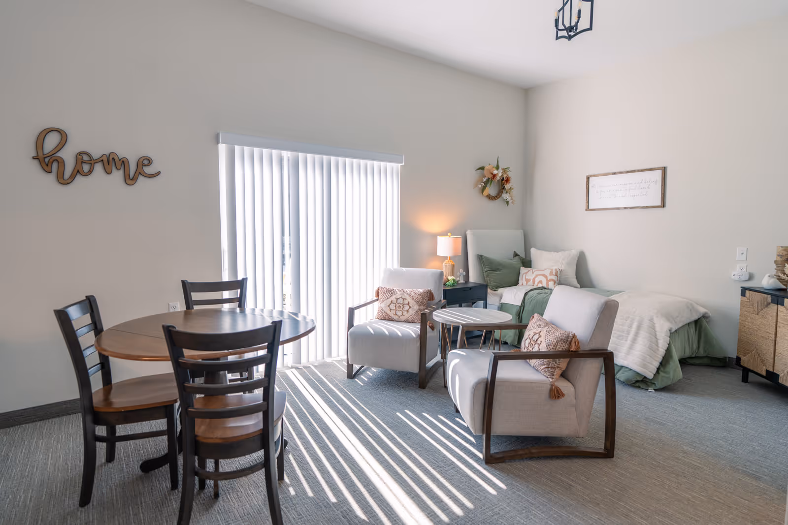 A bright and cozy senior living room with a small round wooden dining table and four chairs on the left. Two cushioned armchairs with decorative pillows face a small round side table. In the background, there is a single bed with green bedding and multiple pillows, a nightstand with a lamp, and wall decorations including a wooden 'home' sign and a floral wreath. Vertical blinds cover a large window letting in sunlight.