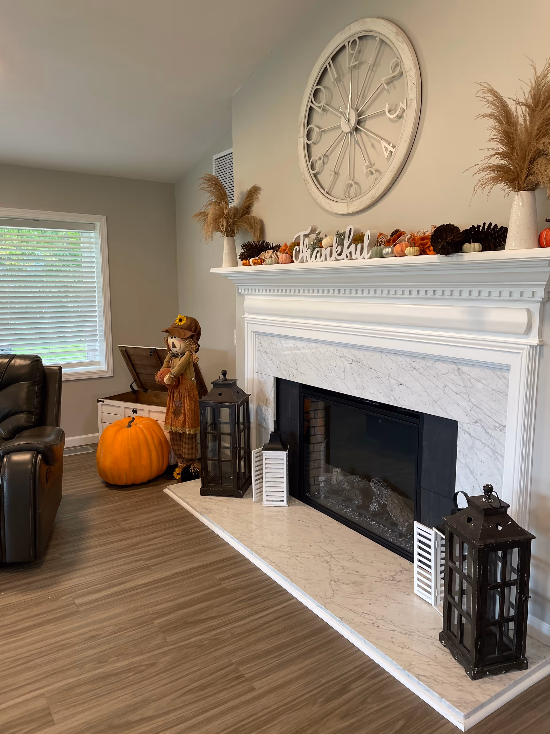Cozy living room featuring a white marble fireplace mantel decorated for fall with pumpkins, lanterns, a large wall clock, and a recliner.