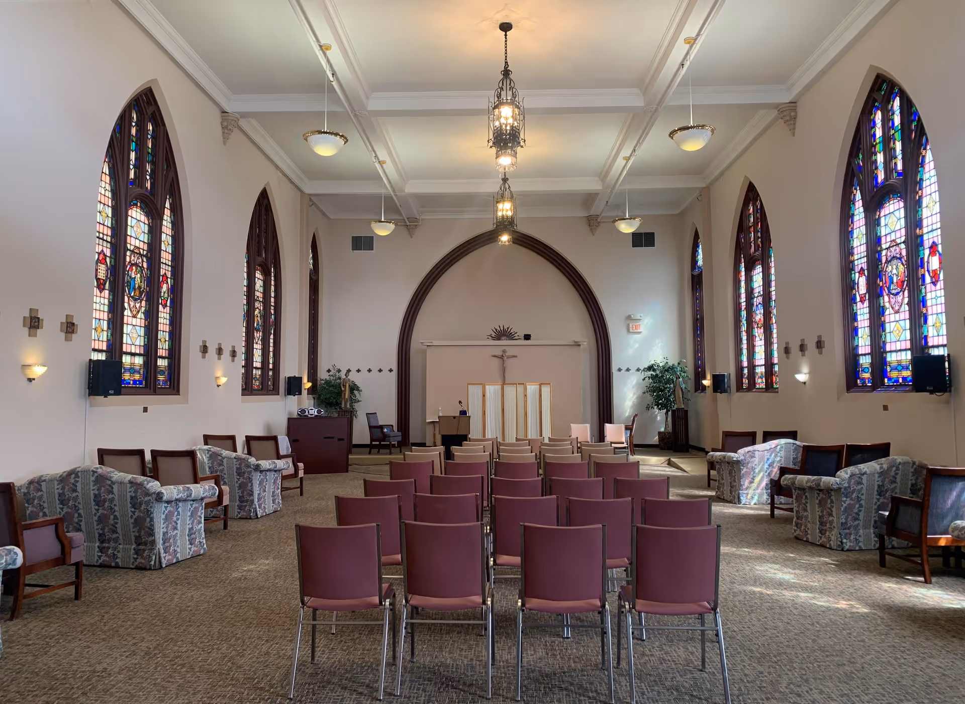 Interior view of a chapel or meeting room with rows of maroon chairs facing a podium and a wooden screen. The room features tall arched stained glass windows on both sides, floral upholstered armchairs along the walls, and hanging light fixtures from a white coffered ceiling.