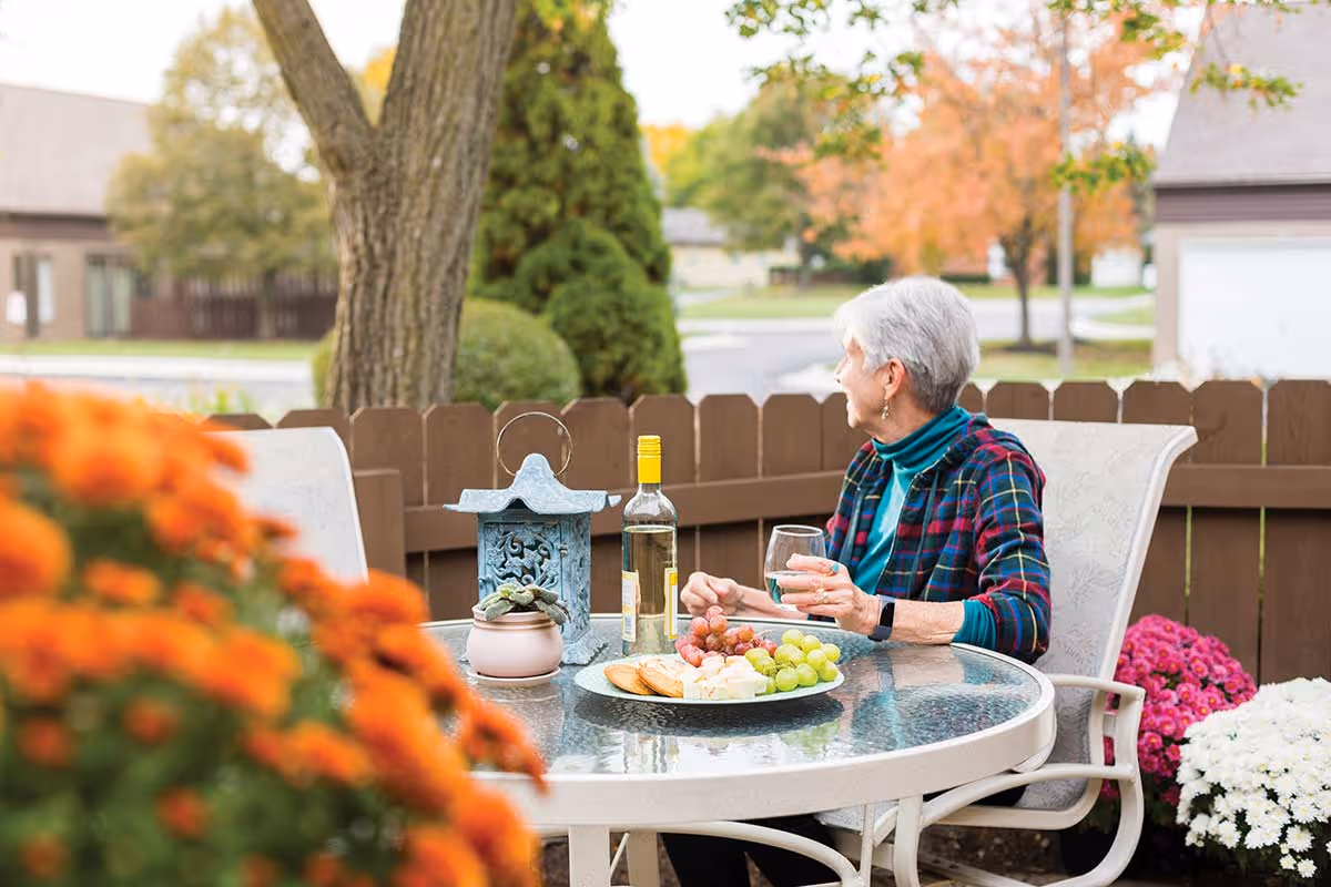 An elderly woman with short gray hair sitting at a round glass outdoor table on a patio, holding a glass of white wine. The table has a plate with grapes, cheese, and crackers, a bottle of white wine, a decorative lantern, and a small potted plant. The patio is surrounded by a wooden fence, with colorful flowers and trees with autumn foliage in the background.