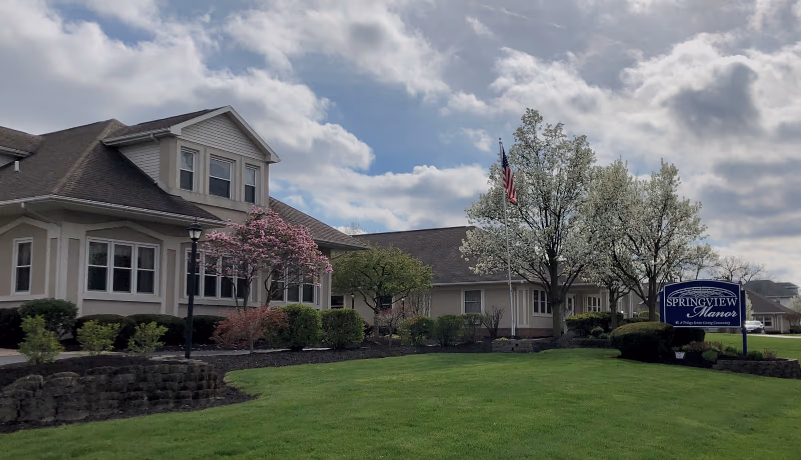 Exterior view of Springview Manor, a senior living facility, showing a beige building with multiple windows, a well-maintained lawn, blooming trees, an American flag on a flagpole, and a blue sign with the facility's name.
