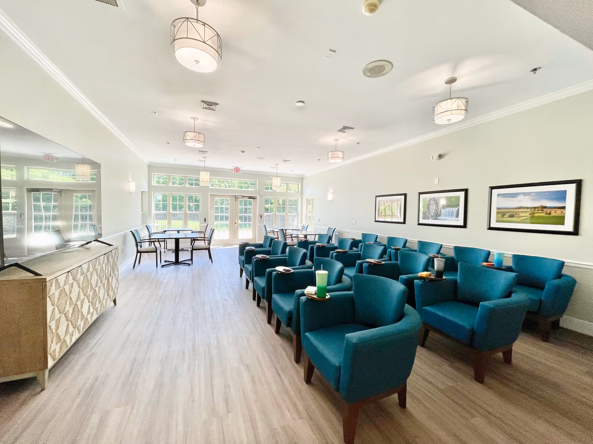 A bright and spacious common area with rows of teal armchairs arranged facing a large flat-screen TV on a wooden cabinet. The room has light-colored walls, wood flooring, and several framed landscape pictures on the wall. Large windows and glass doors at the far end let in natural light, and there are round tables with chairs near the windows.