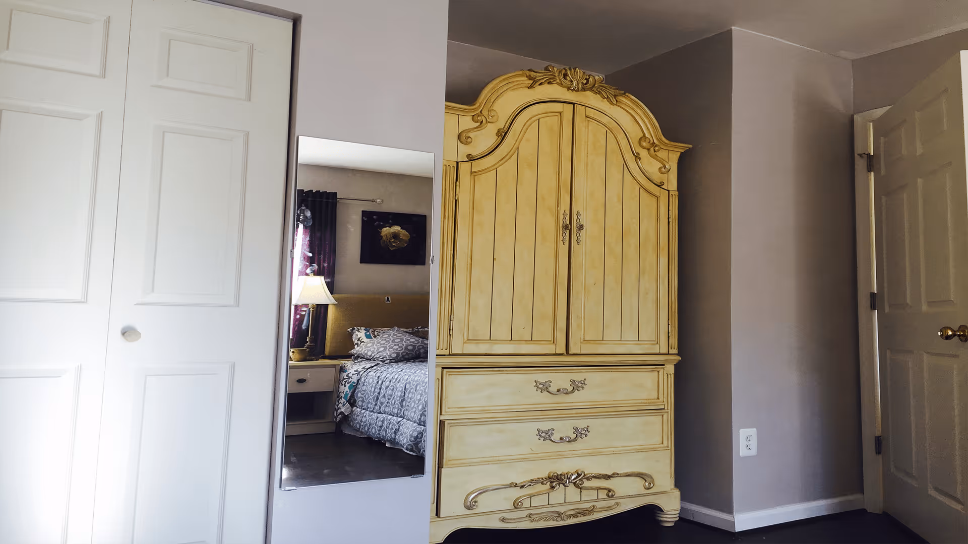 A corner of a bedroom featuring a large ornate yellow armoire with decorative carvings, a white closet door with two panels, and a wall-mounted mirror reflecting a bed with patterned bedding, a nightstand, and a lamp.
