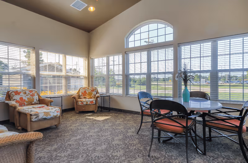 A bright and cozy sitting area with large windows featuring white blinds. The room has two cushioned armchairs with floral upholstery, a round table with four chairs that have orange cushions, and a vase with flowers on the table. The carpet has a subtle floral pattern and the walls are painted light beige.